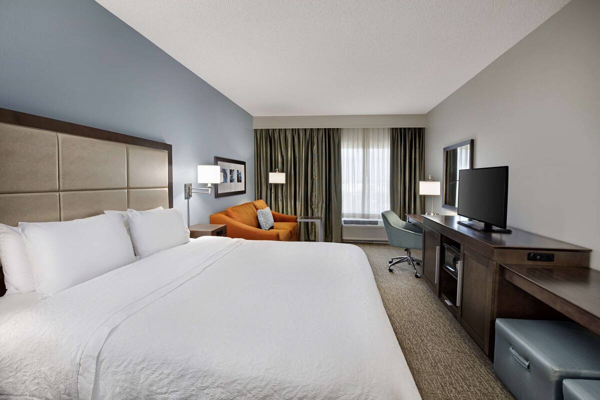 Ein Hotelzimmer mit einem großen Bett, einem Schreibtisch mit Fernseher, einem orangefarbenen Sessel und einem Fenster mit Vorhängen.