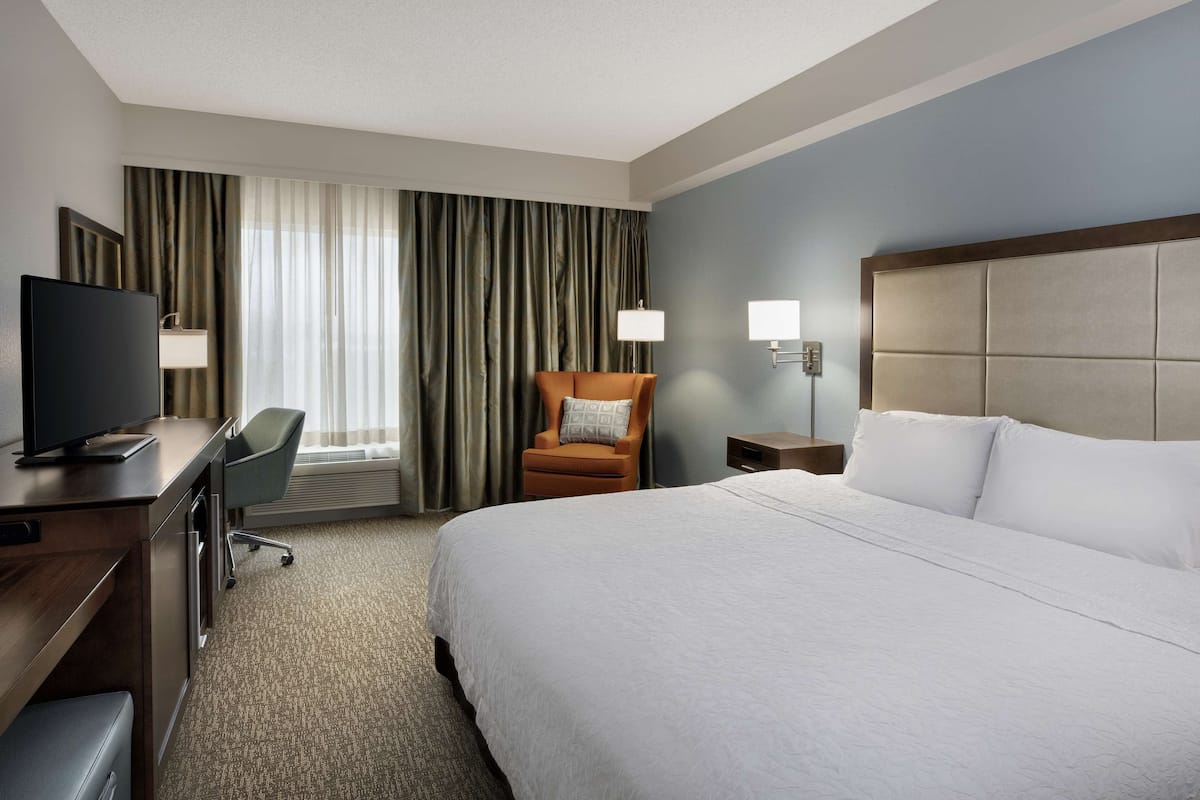 Une chambre d’hôtel avec un grand lit, un bureau avec une télévision, une chaise, une table de chevet et une fenêtre avec des rideaux.