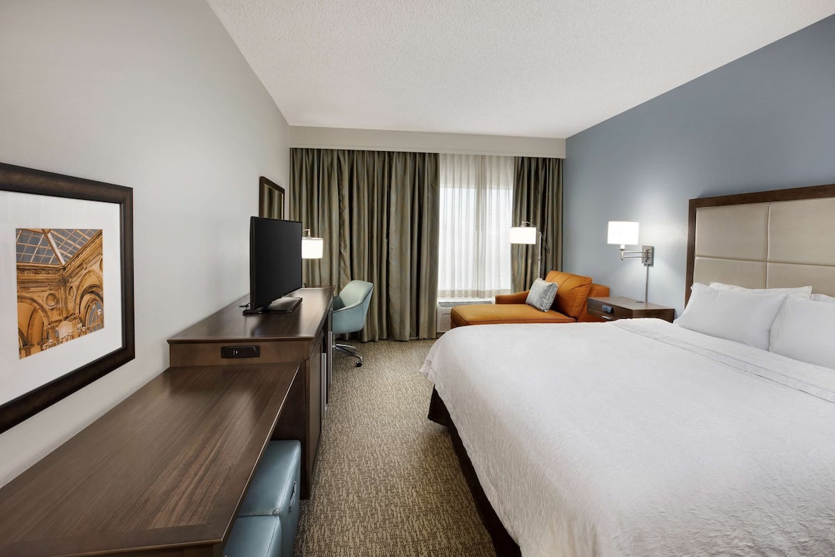 Ein Hotelzimmer mit einem großen Bett, einem Schreibtisch mit Fernseher, einem Sessel und einem Fenster mit Vorhängen.