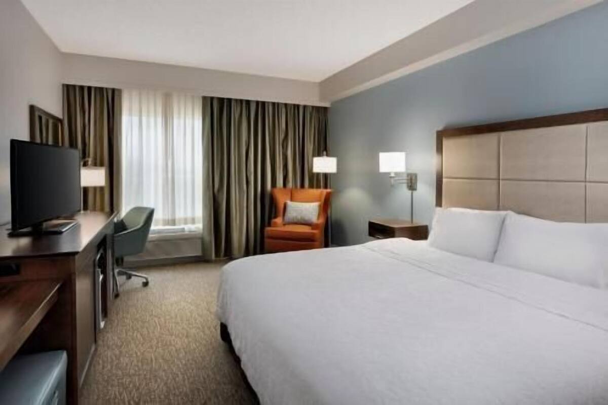 Ein Hotelzimmer mit einem Bett, einem Schreibtisch mit Fernseher, einem Sessel und einem Fenster mit Vorhängen.