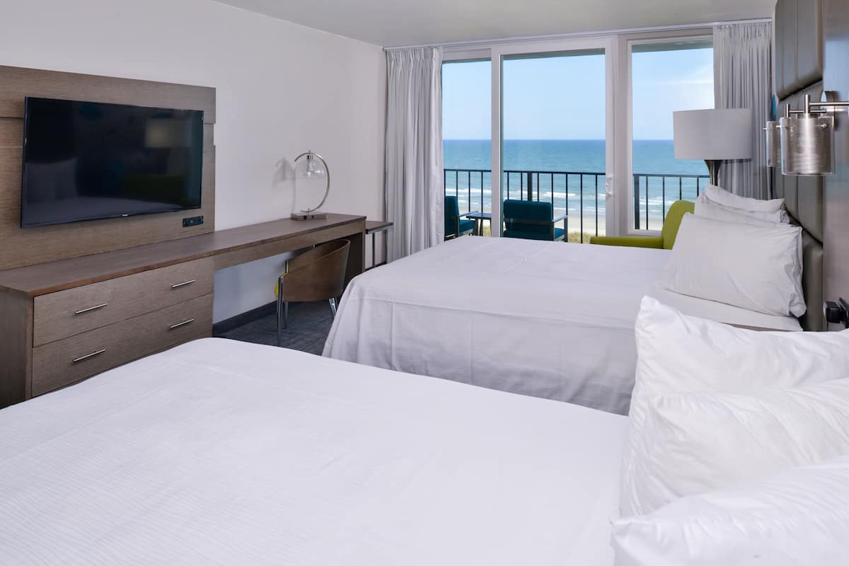 Ett hotellrum med två sängar, ett skrivbord, en TV och en balkong med utsikt över havet.