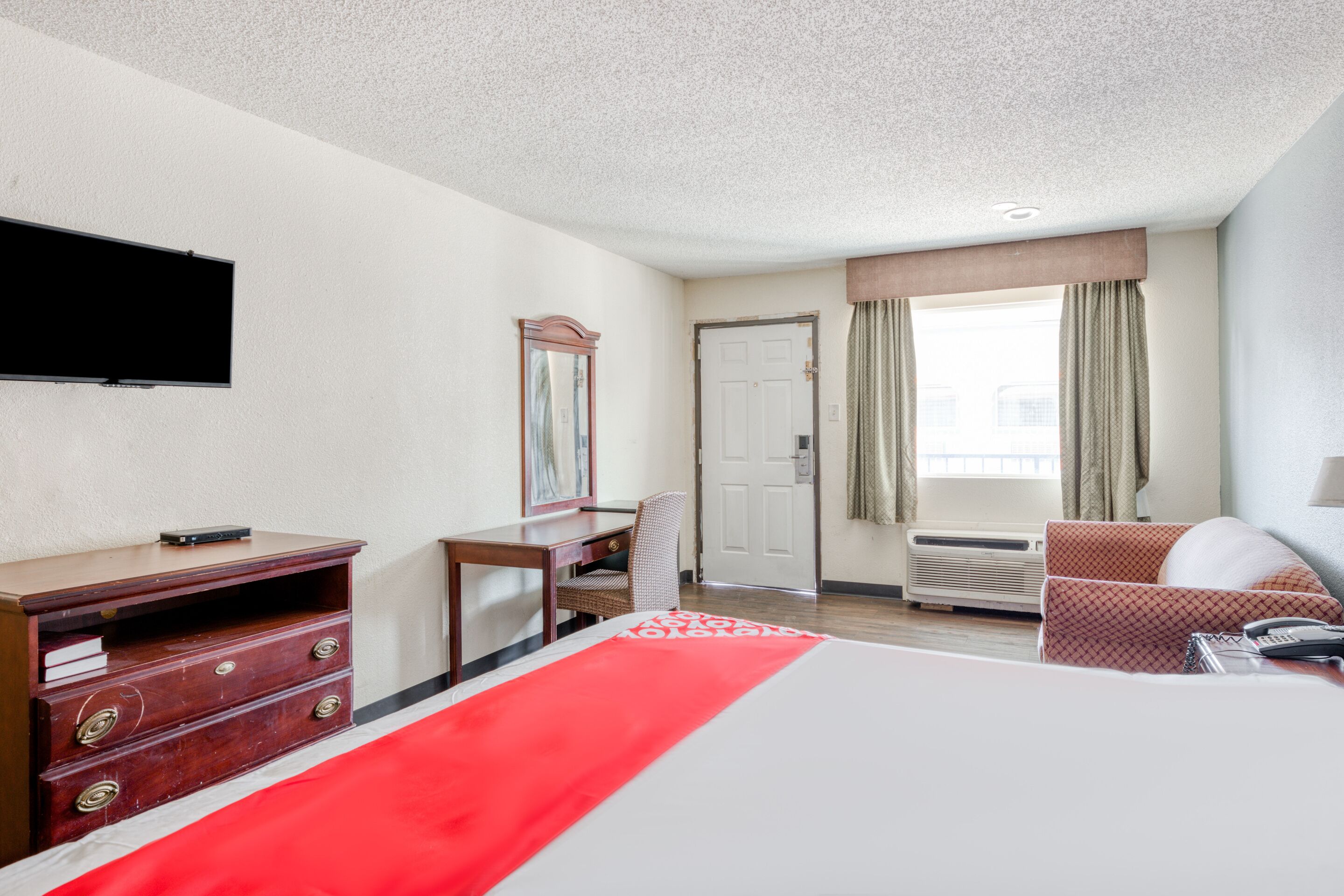 Ein Hotelzimmer mit Bett, Schreibtisch, Stuhl, Kommode, Fernseher, Tür und einem Fenster mit Vorhängen.