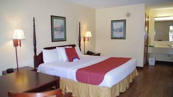 A hotel room with a bed, bedside tables, a desk, and a chair. There are two wall-mounted lamps, a framed picture on the wall, and a mirror above the sink.
