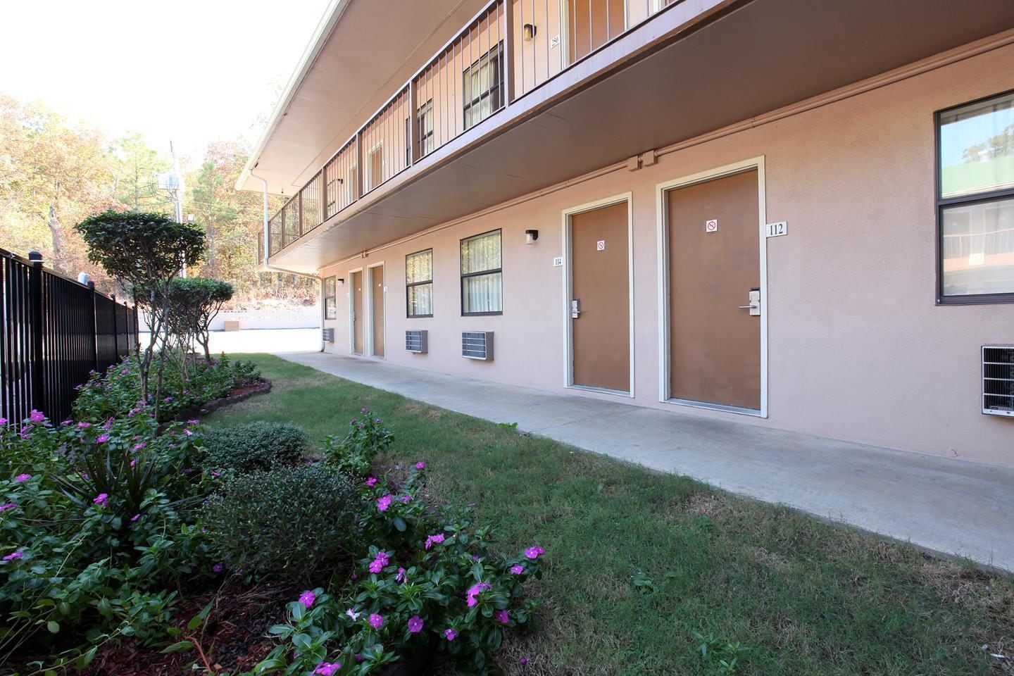 A modern building with a long corridor, numbered doors, and a small garden area with purple flowers.