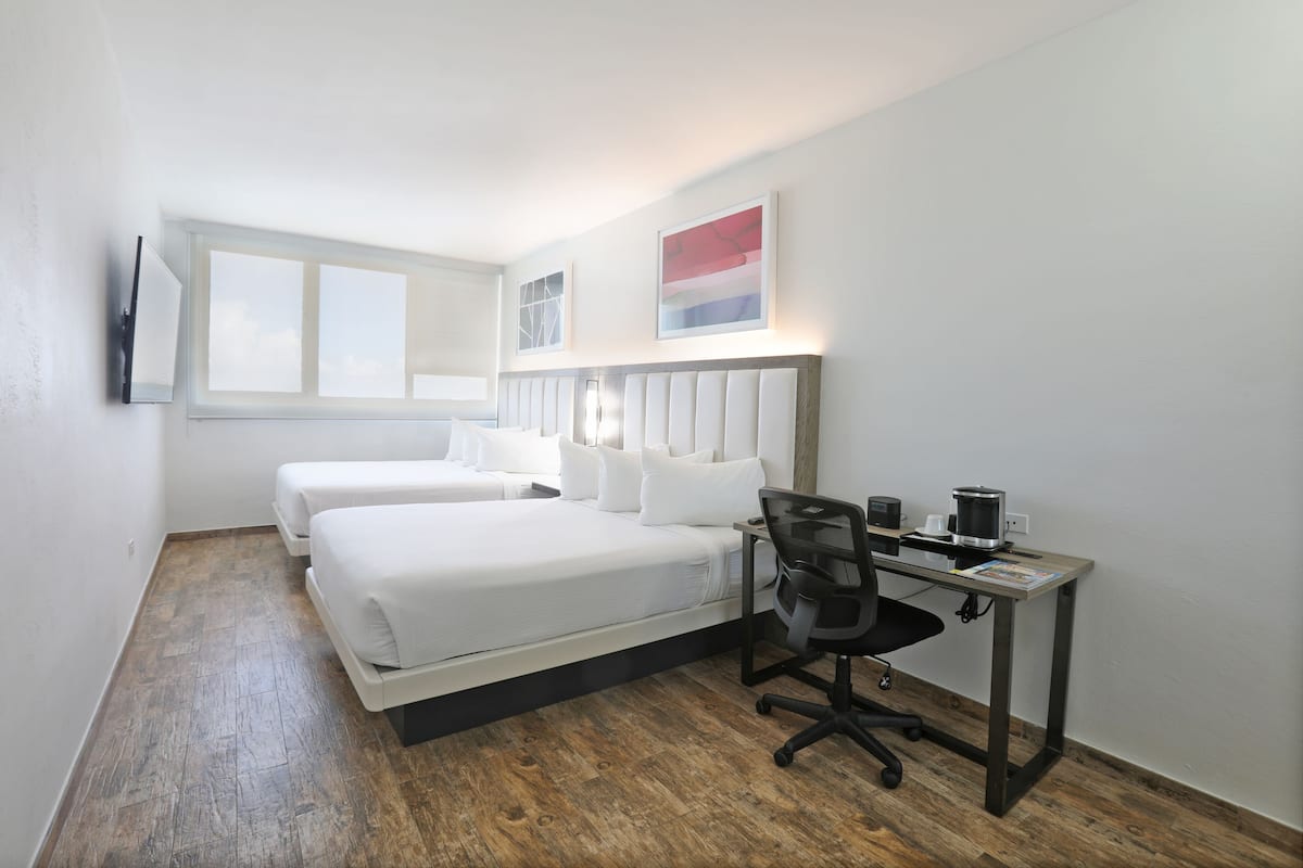 Habitación de hotel con dos camas, un escritorio con silla, cafetera y televisor de pantalla plana montado en la pared.