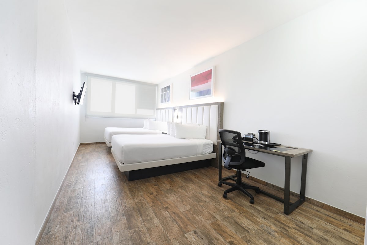 Una habitación pequeña y moderna con suelo de madera, una cama con ropa de cama blanca, un escritorio con una silla y una ventana que proporciona luz natural.