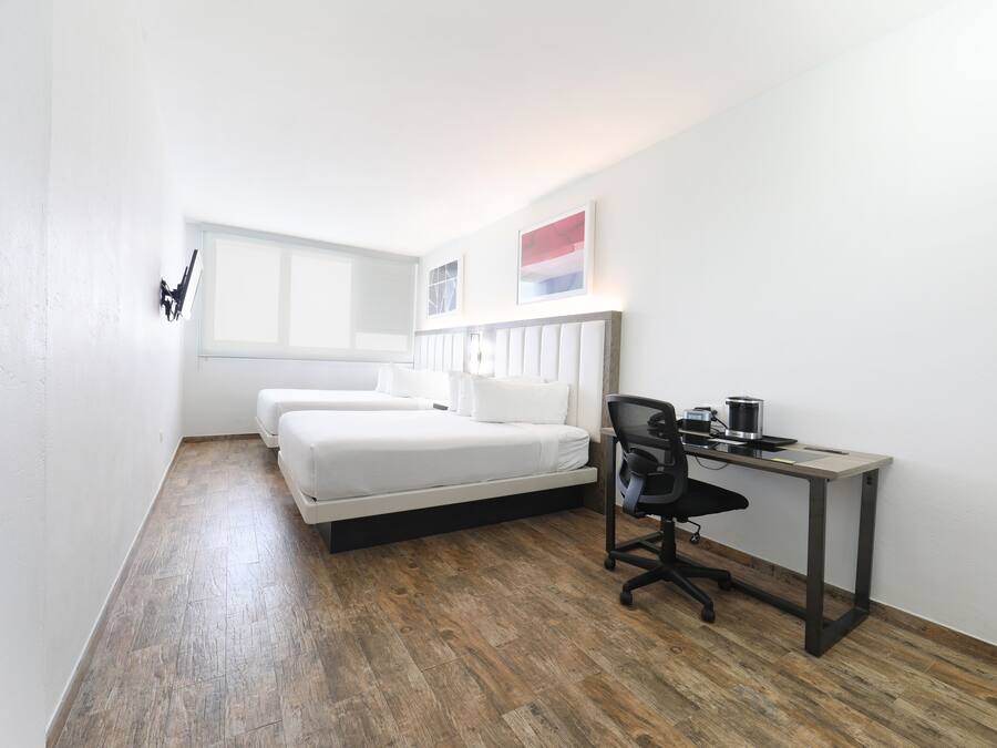 Una habitación pequeña y moderna con suelo de madera, una cama con ropa de cama blanca, un escritorio con una silla y una ventana que proporciona luz natural.