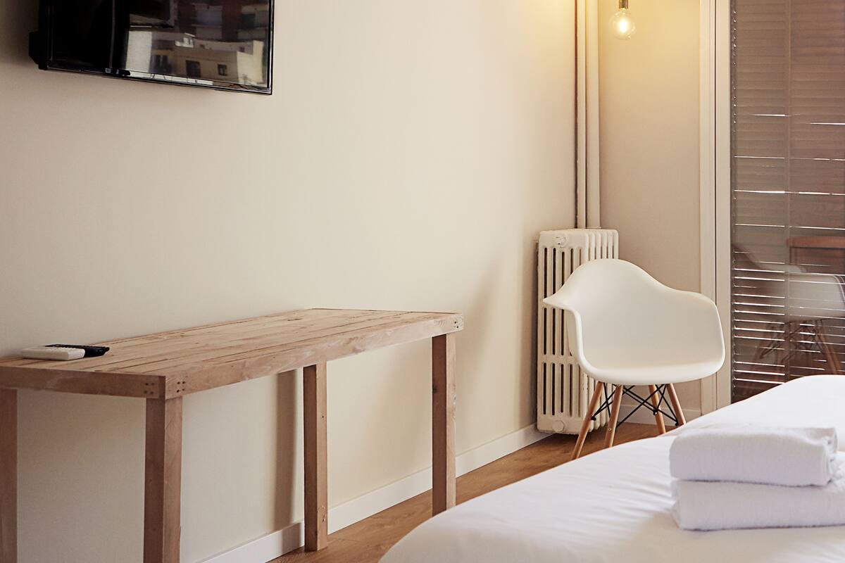 Une chambre d’hôtel avec un lit, une table en bois, une chaise blanche, un radiateur et un téléviseur fixé au mur.