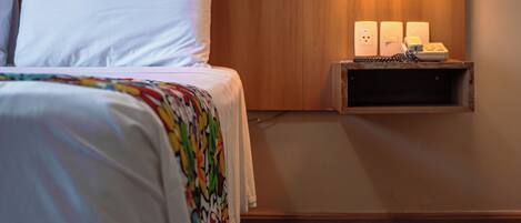 A neatly made bed with white linens, a wooden headboard, and a colorful throw blanket. There is a wall-mounted lamp with three light switches and a small shelf below it.