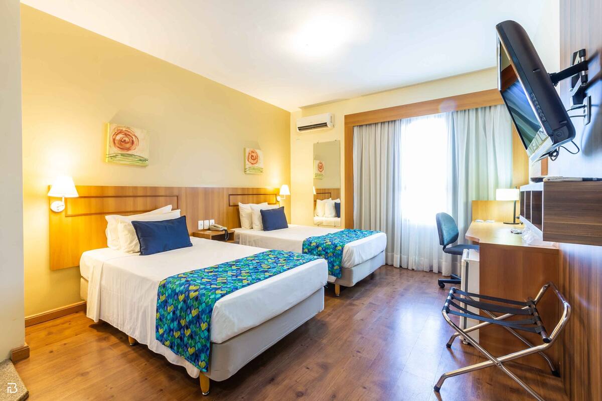 A hotel room with two beds, a desk, a chair, a TV, and artwork on the walls.