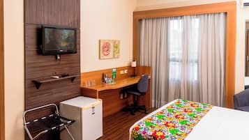 Une chambre d’hôtel équipée d’un lit, d’un bureau, d’une chaise, d’un petit réfrigérateur, d’une télévision, d’œuvres d’art et de rideaux.