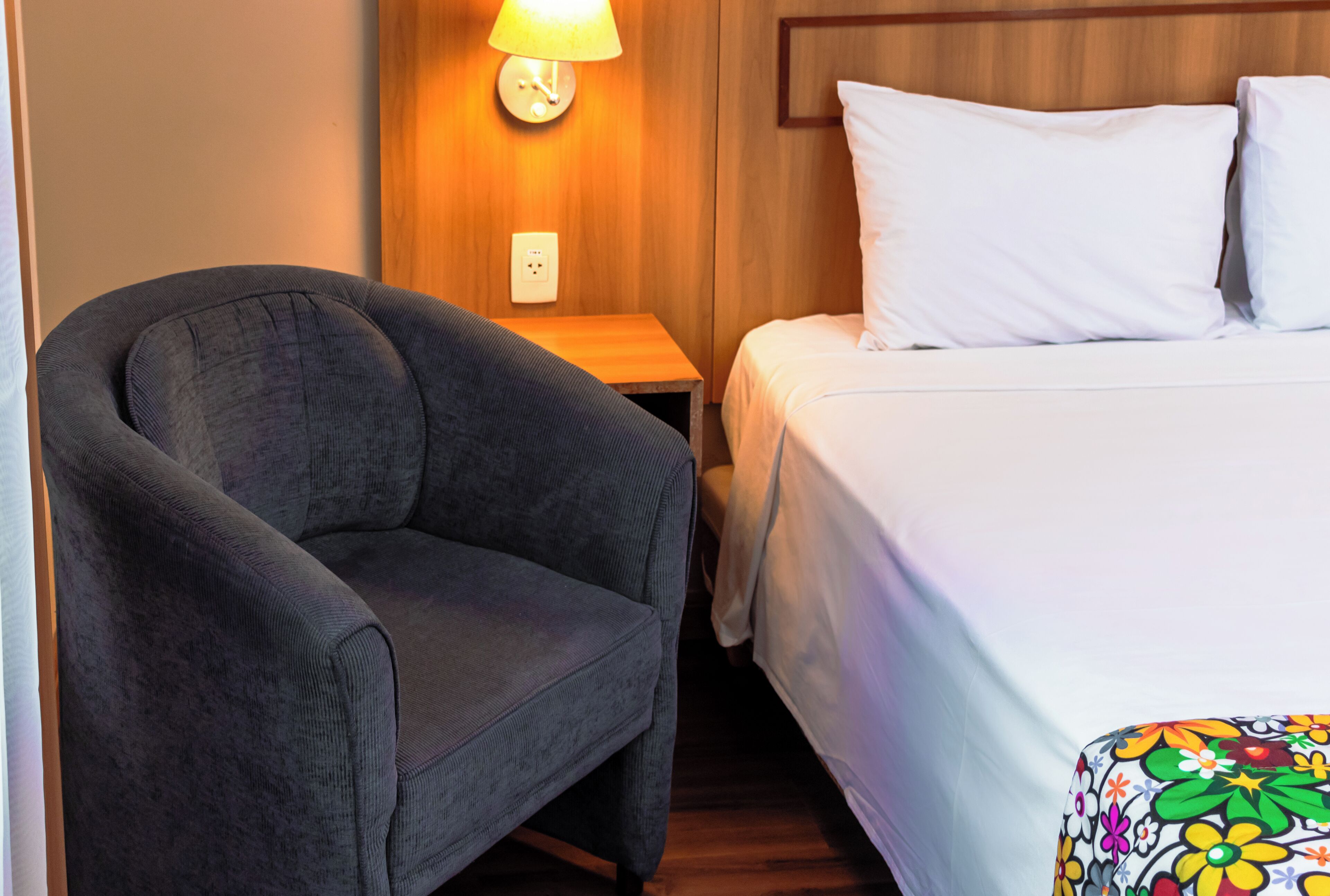 Una habitación de hotel con un sillón gris, una mesita de noche de madera con una lámpara, y una cama con ropa de cama blanca y un plaid floral colorido.