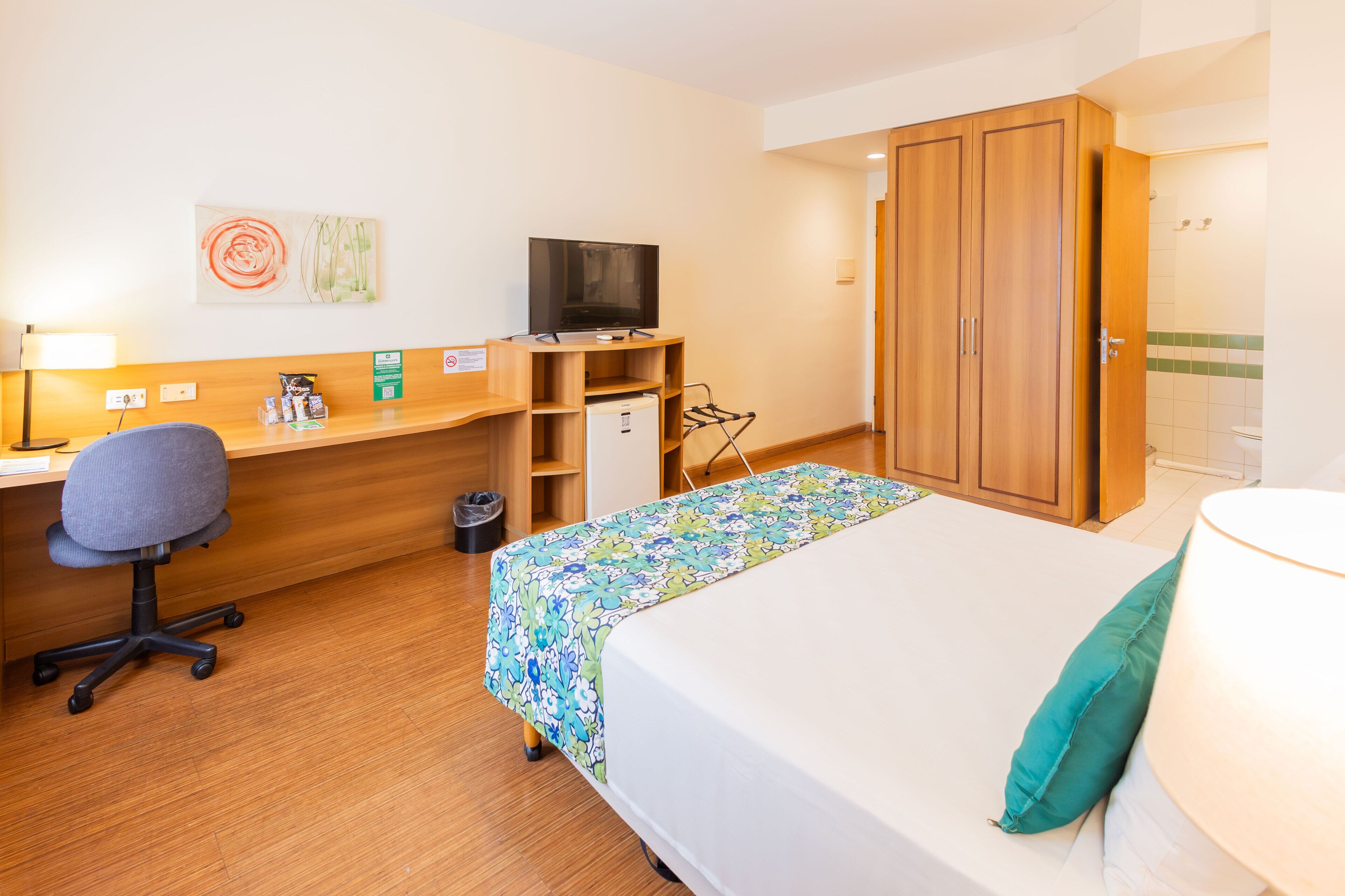 A hotel room with a bed, desk, chair, TV, wardrobe, and a small fridge.