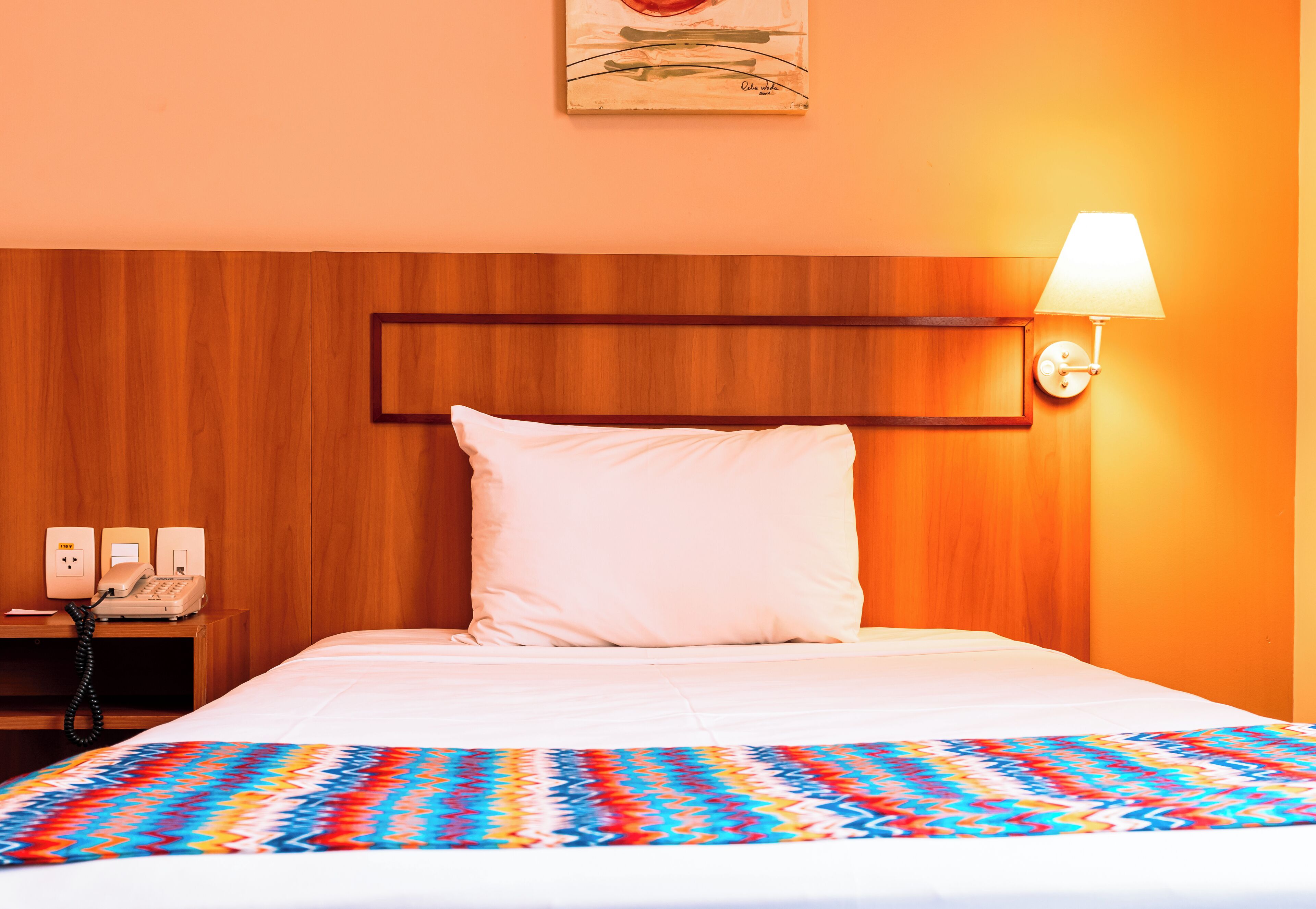 Quarto de hotel com cabeceira de madeira, travesseiro branco, manta colorida, abajur e telefone em uma mesinha de cabeceira.