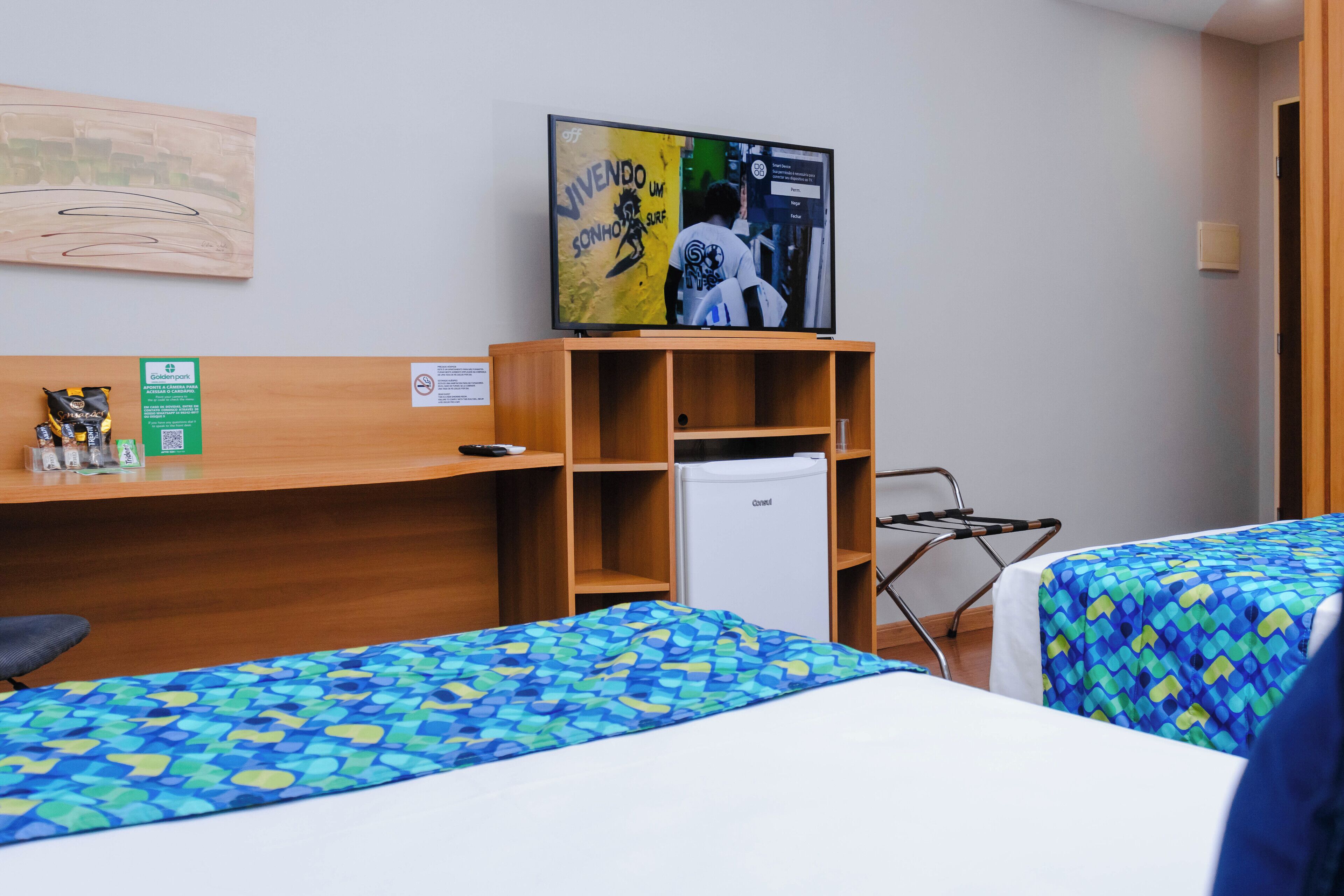 A hotel room with two beds, a television, a small refrigerator, and a wooden shelf.