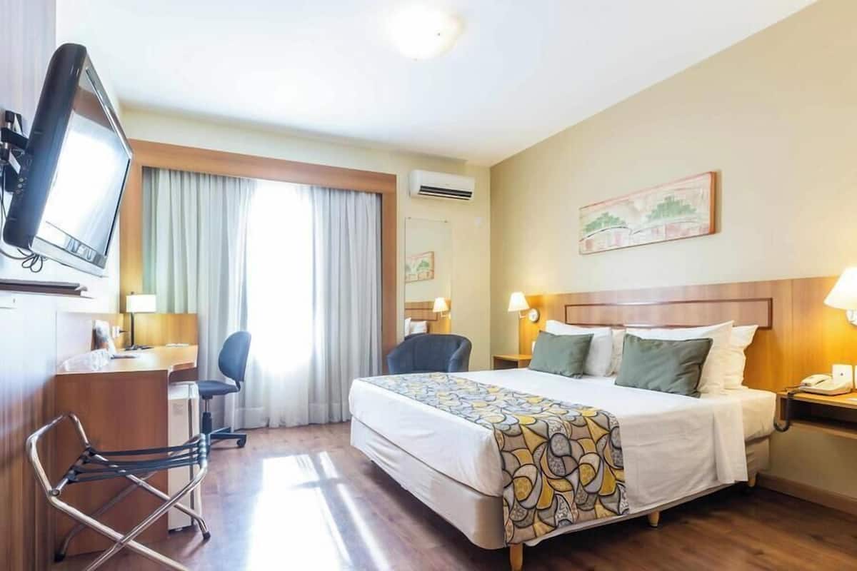 A hotel room with a bed, desk, chair, TV, and artwork on the wall.