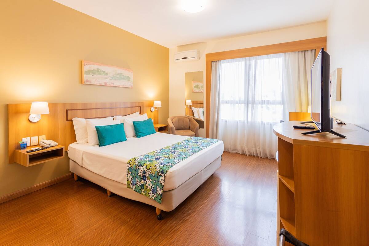 A hotel room with a bed, bedside tables with lamps, a desk with a TV, and a window with curtains.