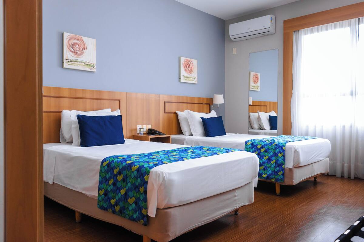 A hotel room with two beds, each with a blue and yellow patterned bedspread, white linens, and a wooden headboard. There is a nightstand with a lamp and a phone, and a window with white curtains.