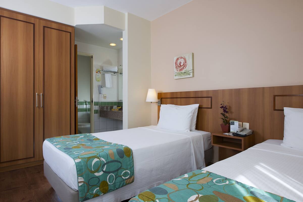 A hotel room with two beds, a wooden wardrobe, a bathroom visible through an open door, and a wall-mounted artwork.