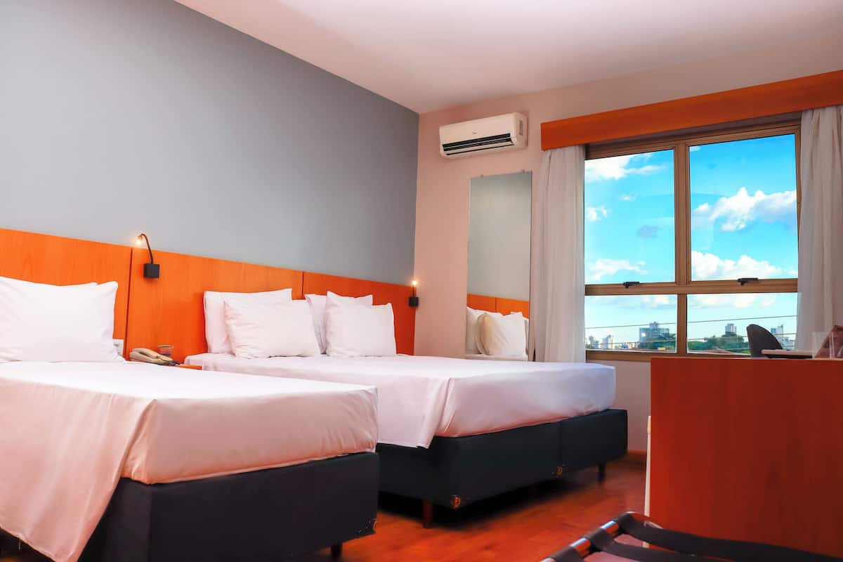 A hotel room with two beds, a desk, and a large window with a city view.
