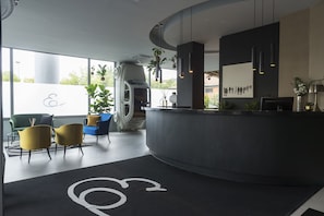 A modern reception area with a curved desk, seating with cushions, and a large window offering a view of greenery. - Elizabeth Lifestyle Hotel (Bologna)