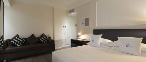 A hotel room with a large bed, a dark brown sofa with patterned cushions, a bedside table with a lamp, and a mirror on the wall.