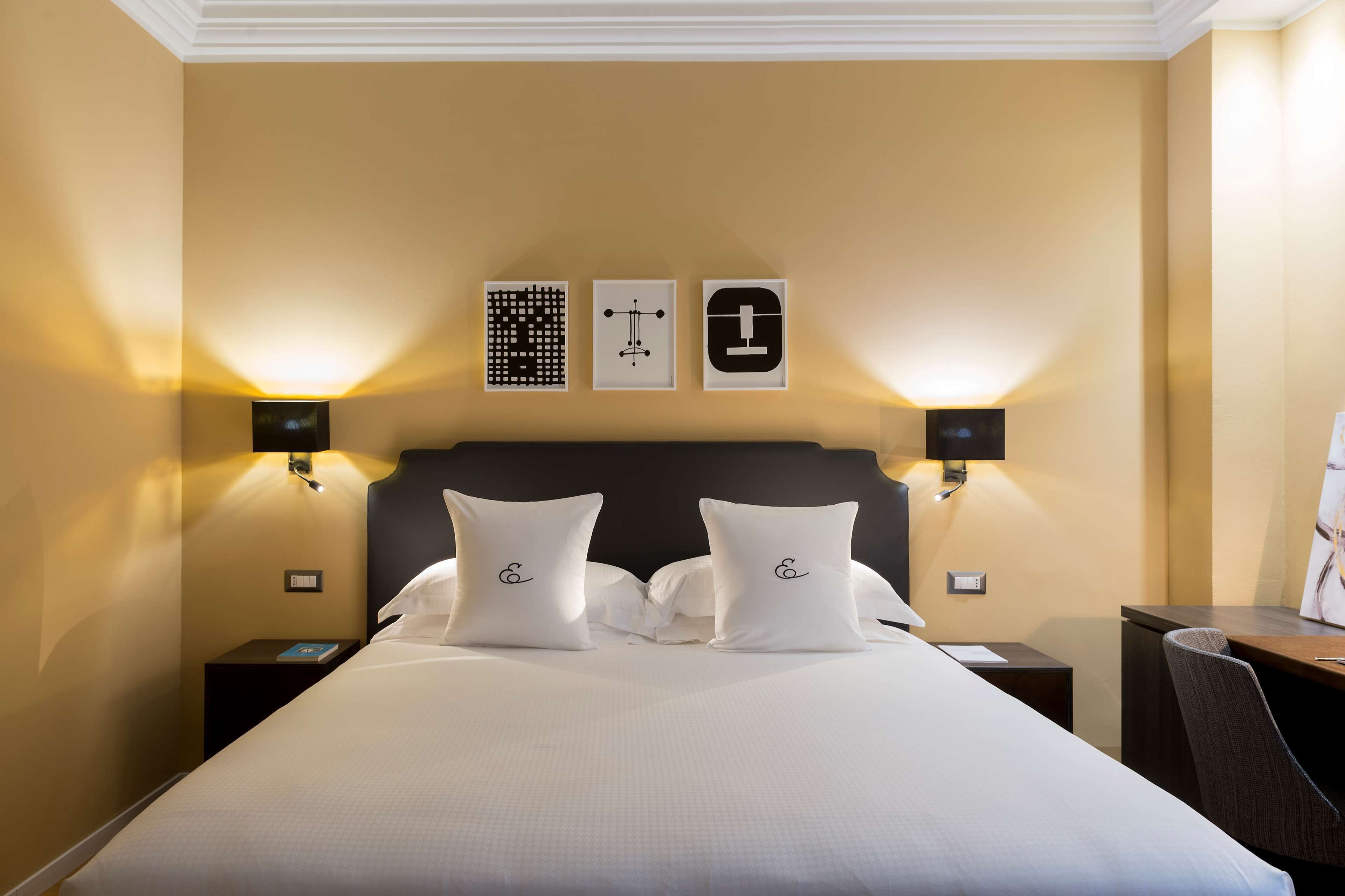 Ein Hotelzimmer mit einem großen Bett, zwei Nachttischen, einem Schreibtisch und einem Sessel. An den Wänden sind Leuchten angebracht und über dem Bett hängen drei gerahmte Bilder.