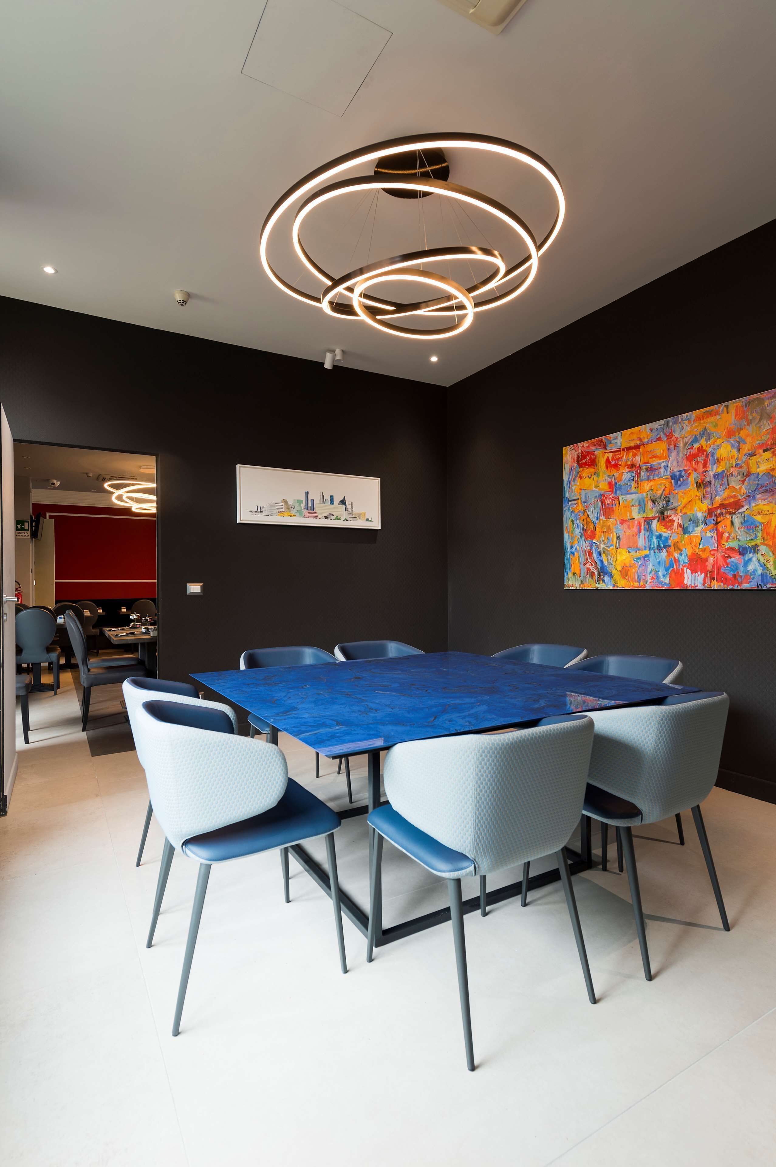 Een moderne eethoek met een blauwe tafel en grijze stoelen, een plafondlamp en een kleurrijk abstract schilderij aan de muur.