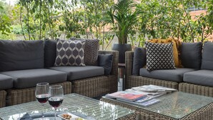 A covered outdoor seating area with wicker furniture, a glass-top table with drinks, and a view of greenery and buildings in the background. - Elizabeth Lifestyle Hotel (Bologna)