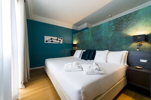 Hotelkamer met een groot bed, witte beddengoed, teal muren, een modern hoofdeinde en een nachtkastje met een lamp.