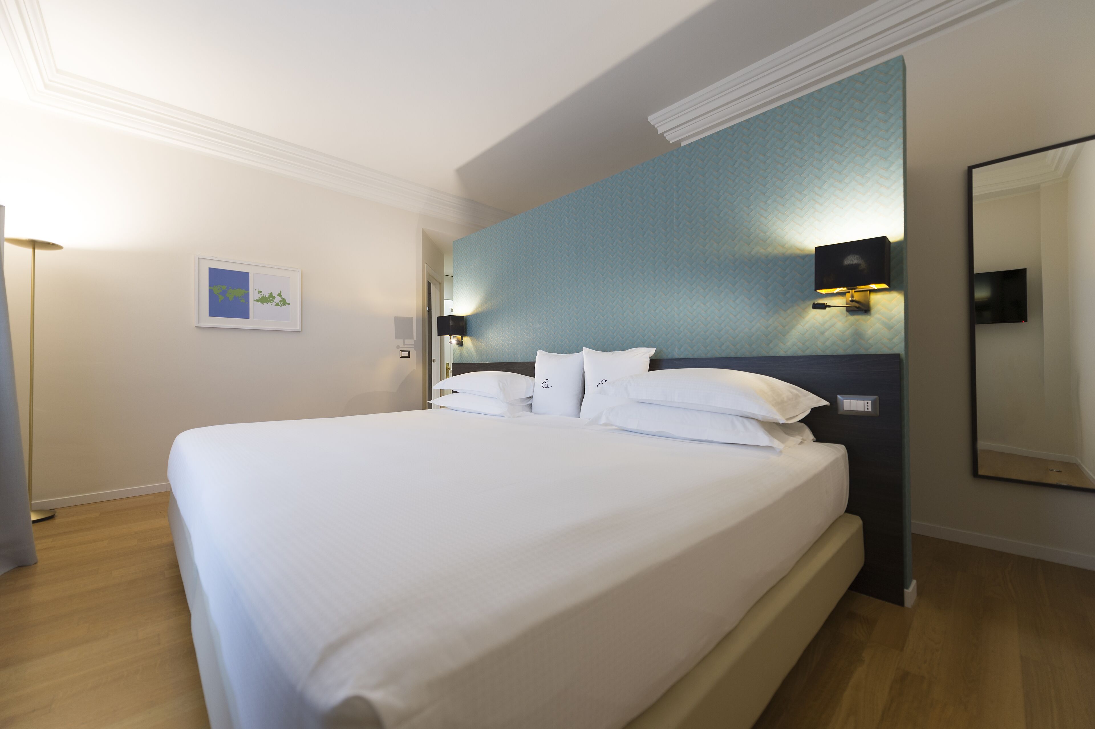 A neatly made bed in a hotel room with a large mirror, a wall-mounted TV, and a framed picture on the wall.