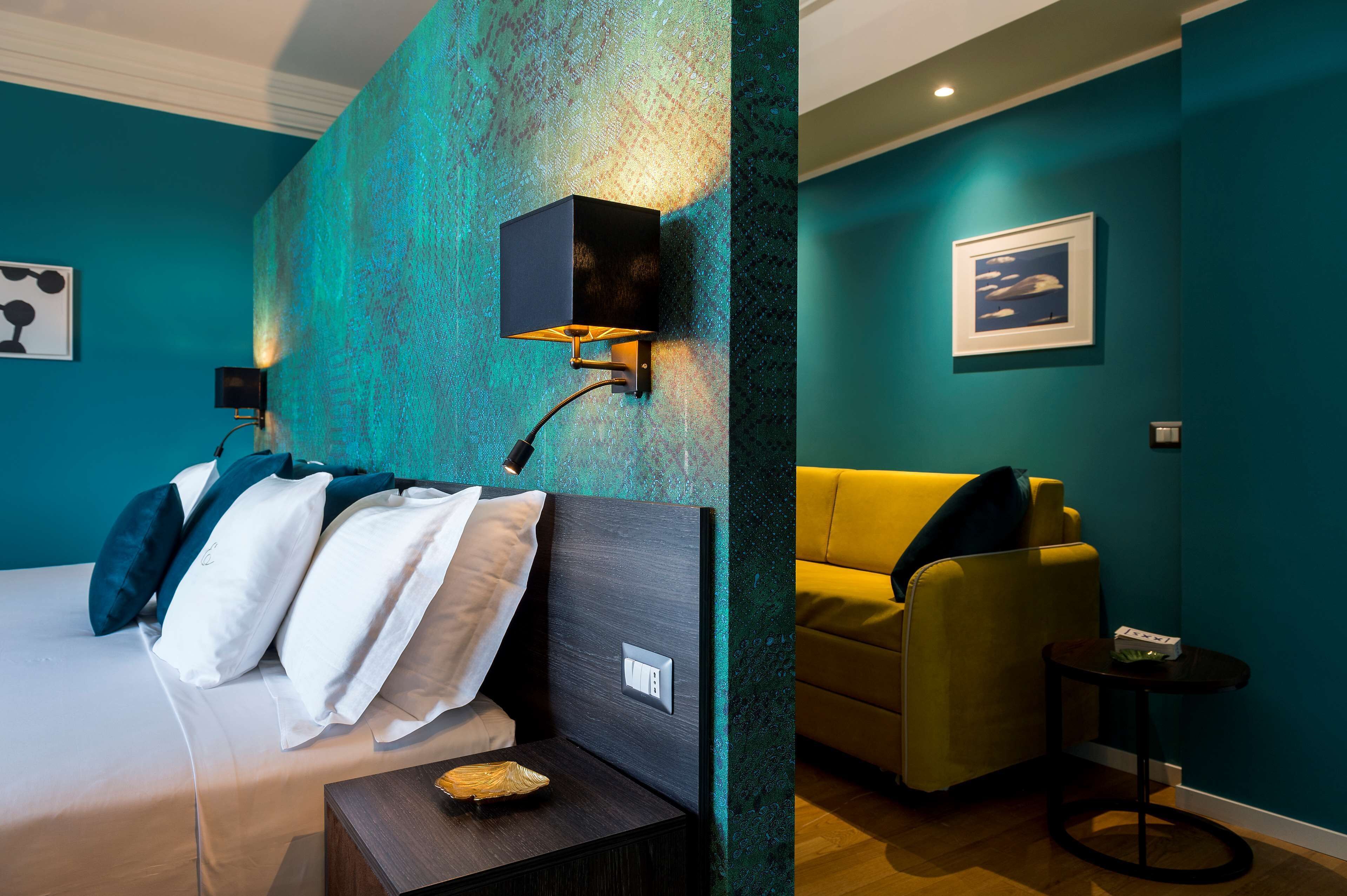 A modern hotel room with teal walls, a bed with white and teal bedding, a yellow armchair, and a small side table.