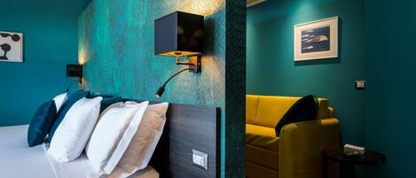 A modern hotel room with teal walls, a bed with white and teal bedding, a yellow armchair, and a small side table.