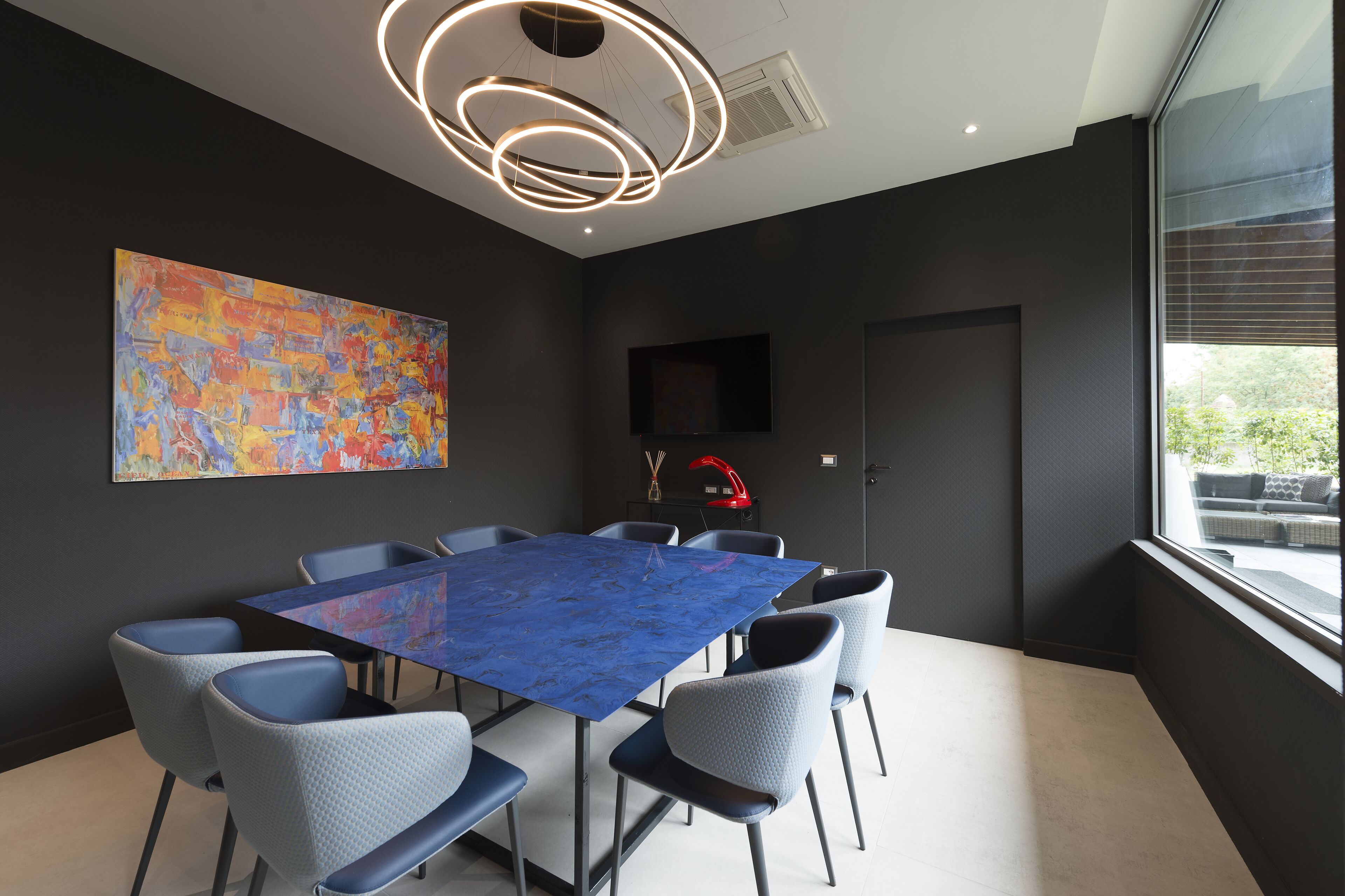 A modern meeting room with a blue table, grey chairs, a large abstract painting, a red lamp, and a window with a view.