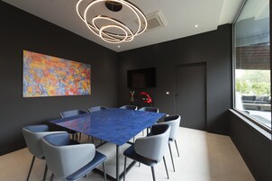 Een moderne vergaderruimte met een blauwe tafel, grijze stoelen, een groot abstract schilderij, een rode lamp en een raam met uitzicht.