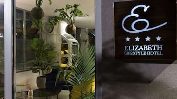 Une enseigne indiquant l’hôtel Elizabeth Lifestyle, dont on aperçoit l’intérieur moderne à travers une fenêtre, avec des plantes en pot et des meubles contemporains.
