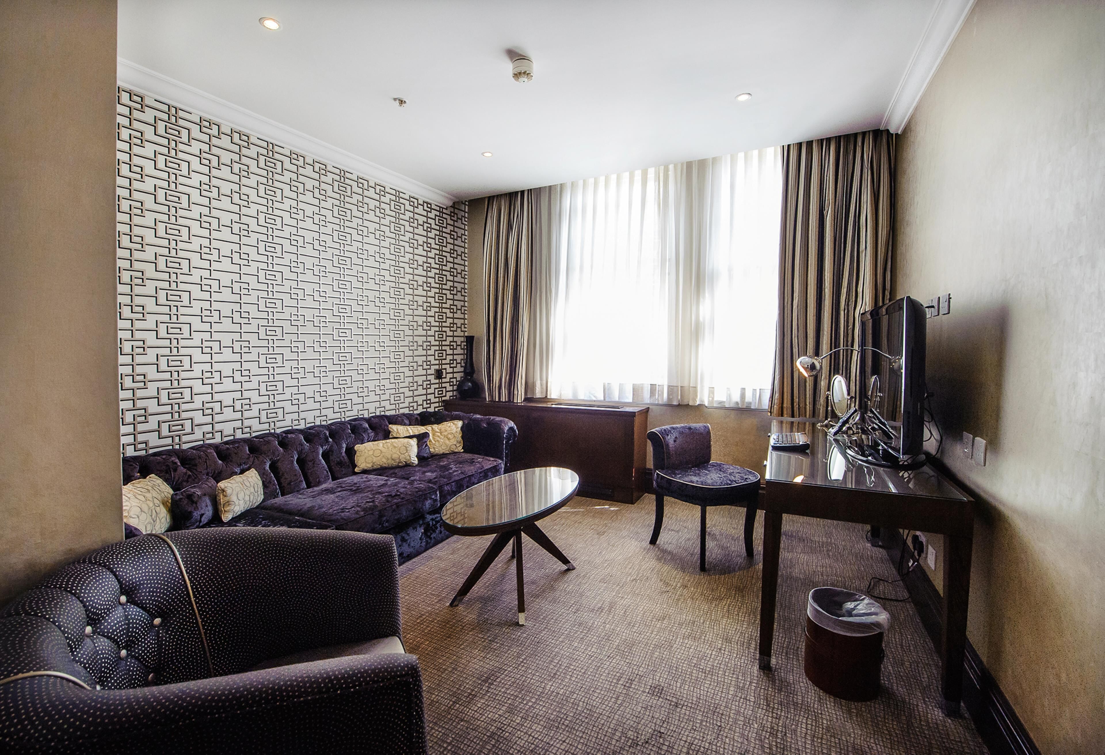 A hotel room with a patterned wall, a sofa, a small table, a chair, a television, and a window with curtains.