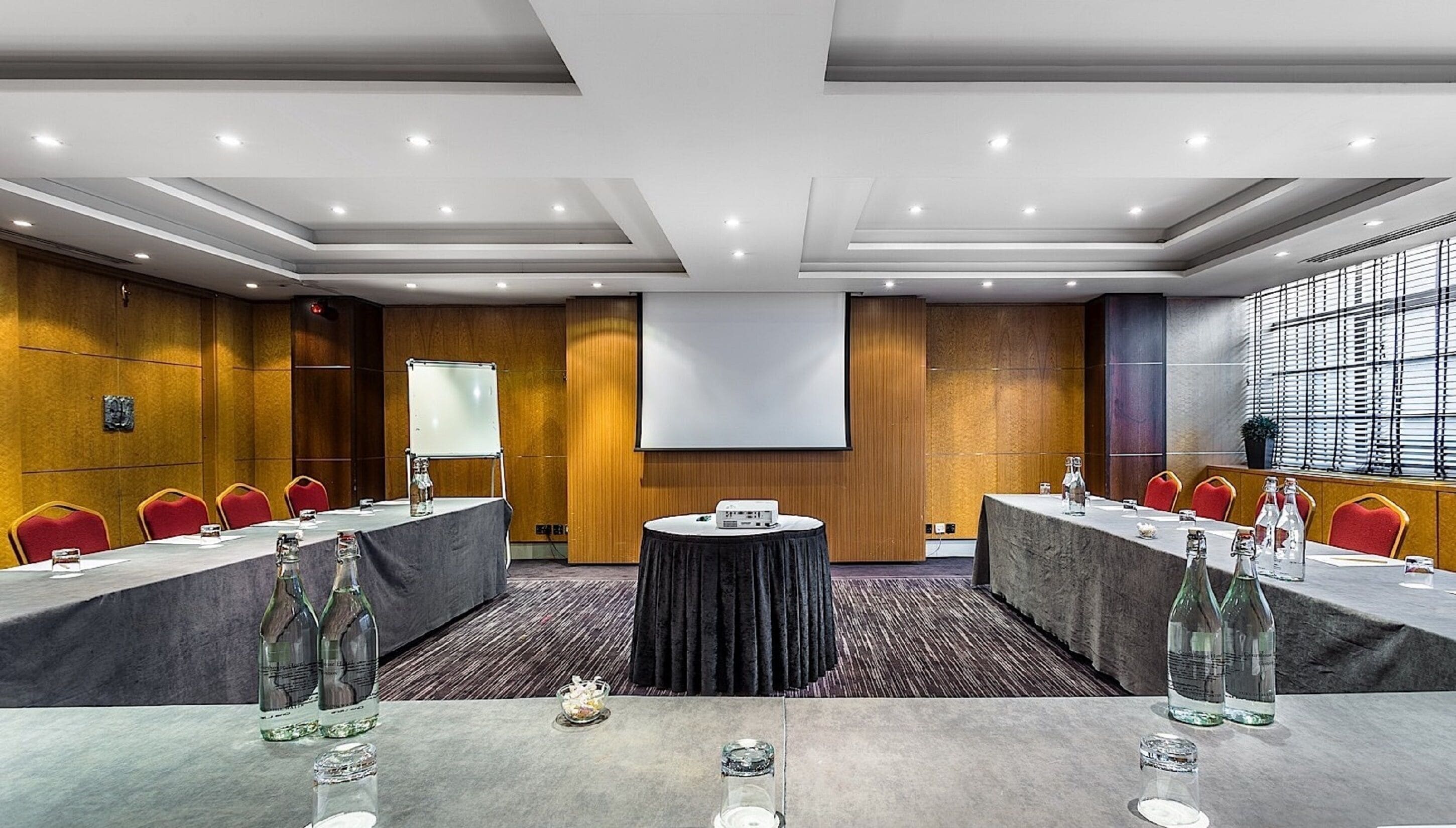 A conference room with a long table, red chairs, a projector screen, and a large window with blinds.