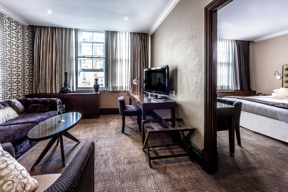 Ein Hotelzimmer mit einer Couch, einem Glastisch, einem Schreibtisch mit Fernseher, einem Sessel, einem Bett und einem großen Fenster mit Vorhängen.