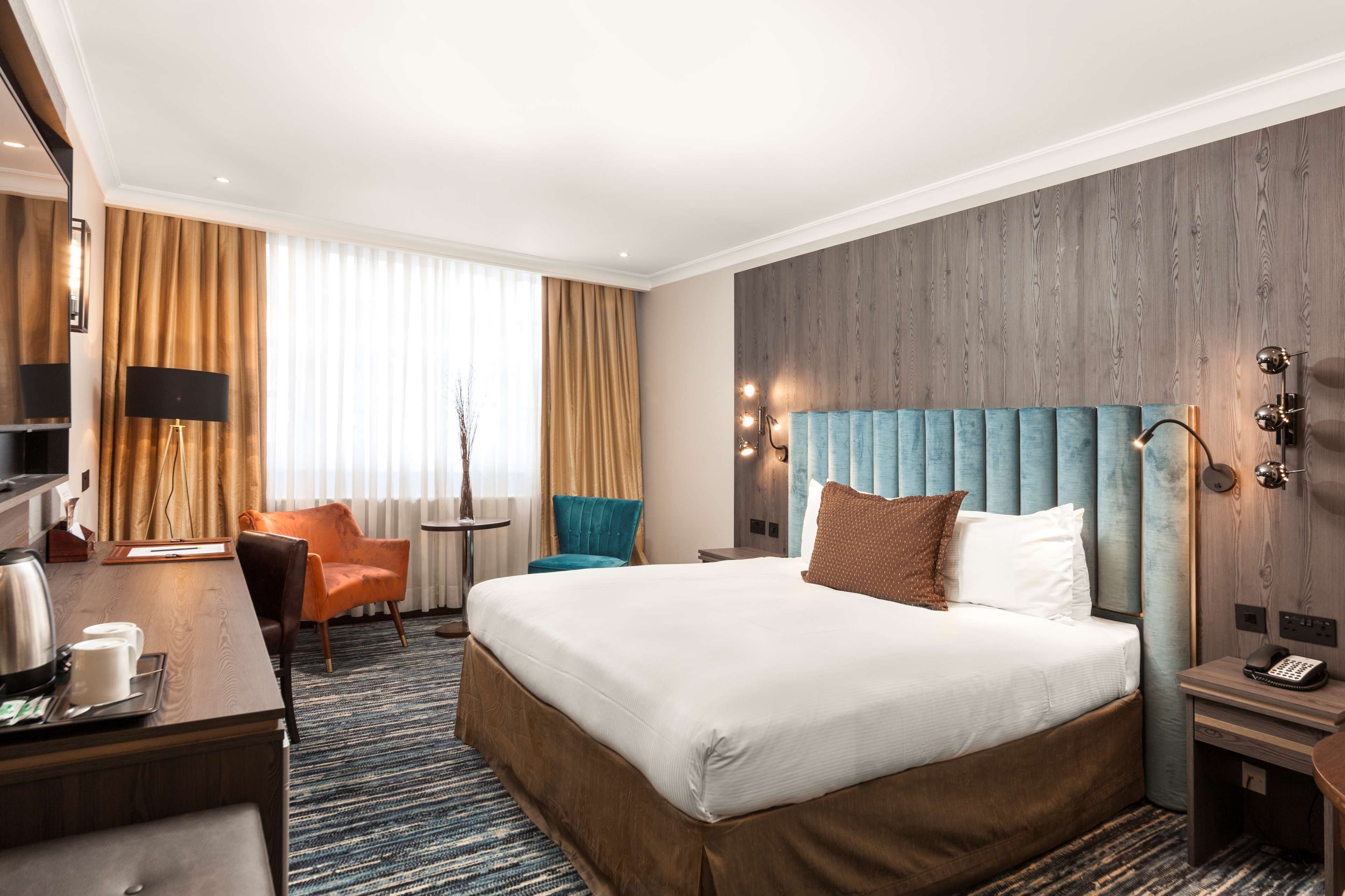 Une chambre d’hôtel avec un grand lit, un bureau avec une machine à café, une chaise, une petite table et une lampe.