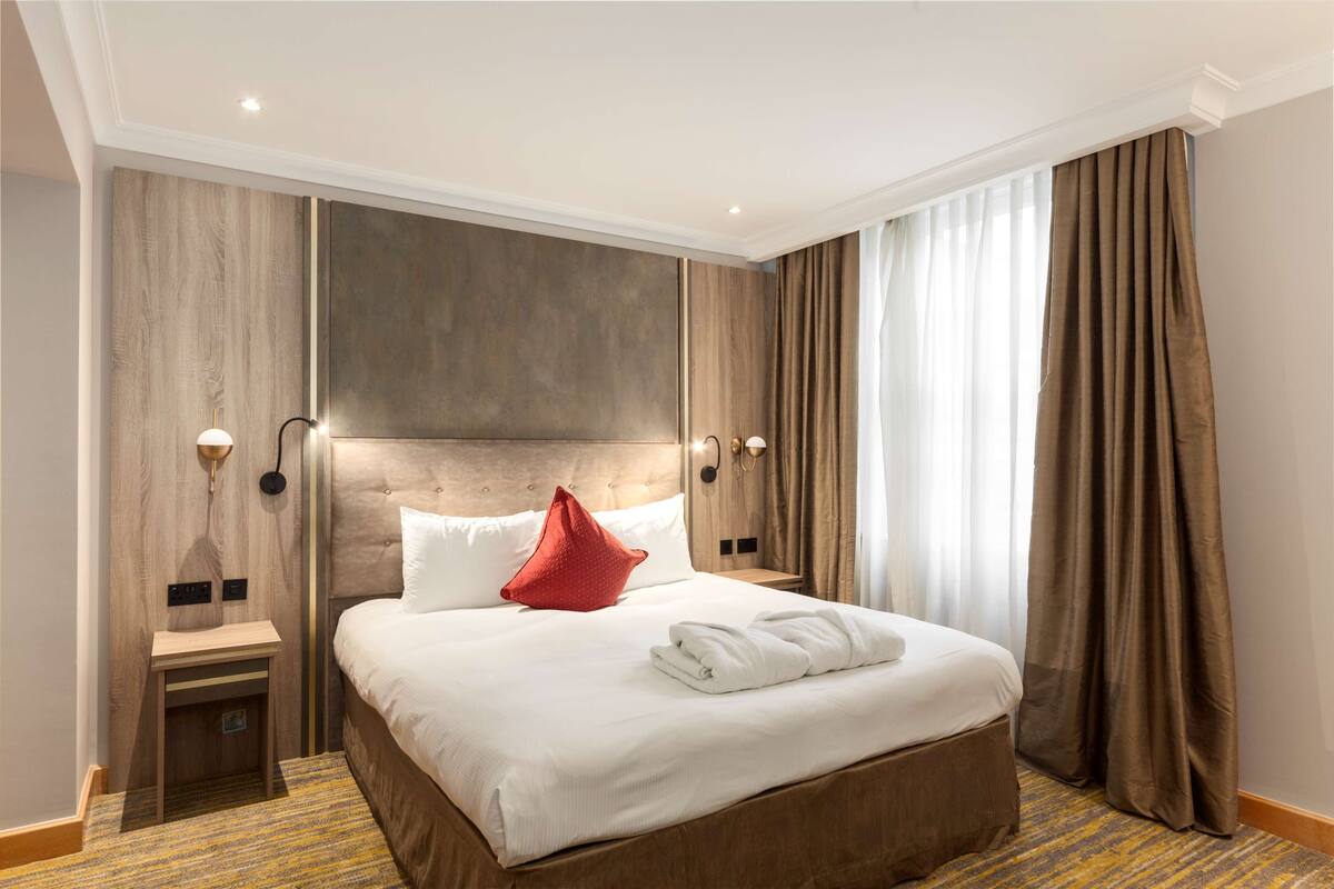 Ein Hotelzimmer mit einem großen Bett, einem Nachttisch, einer Wandlampe und einem Fenster mit Vorhängen.