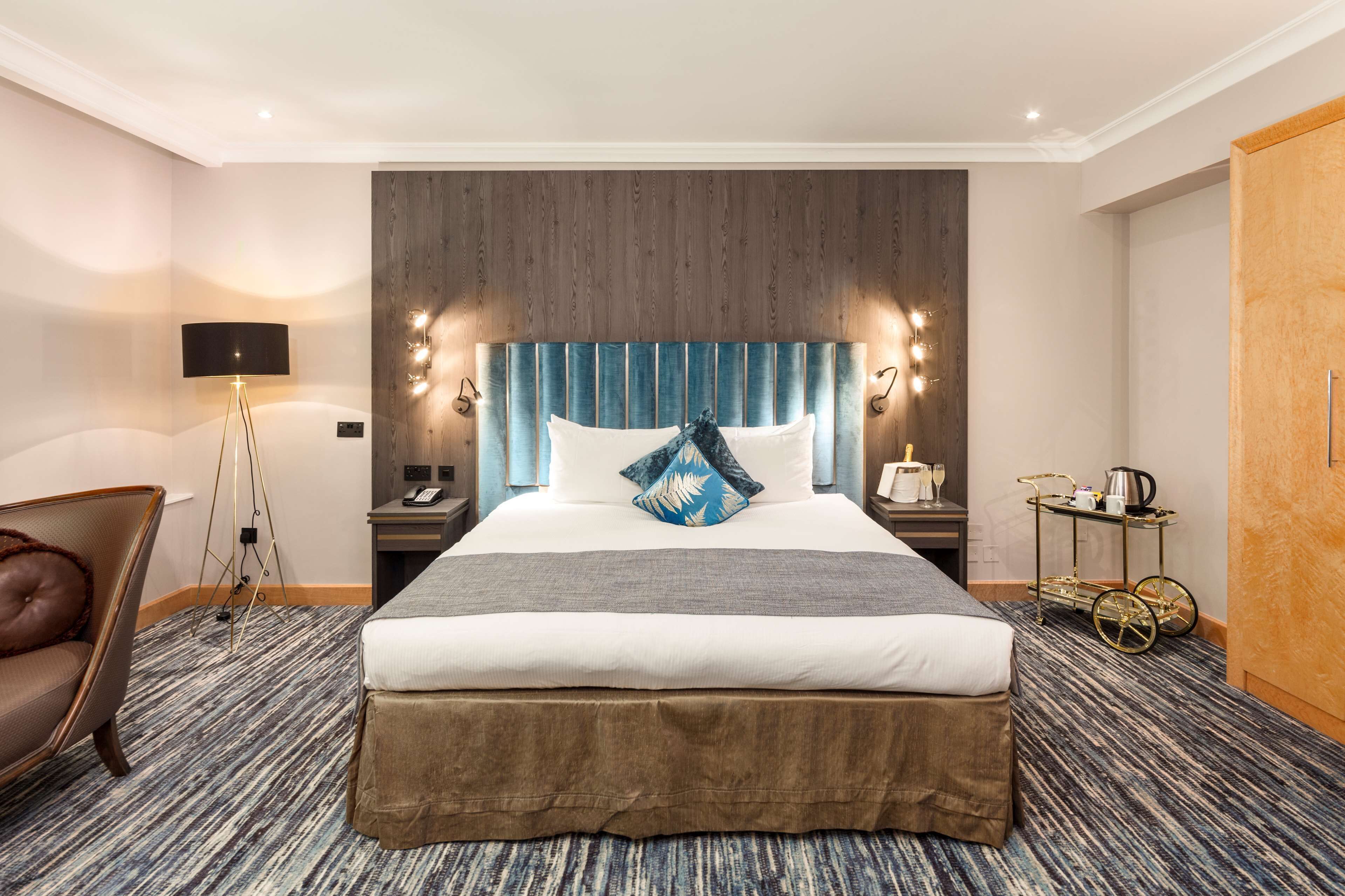 A hotel room with a large bed, a chair, a bedside table with a lamp, and a small table with a kettle.