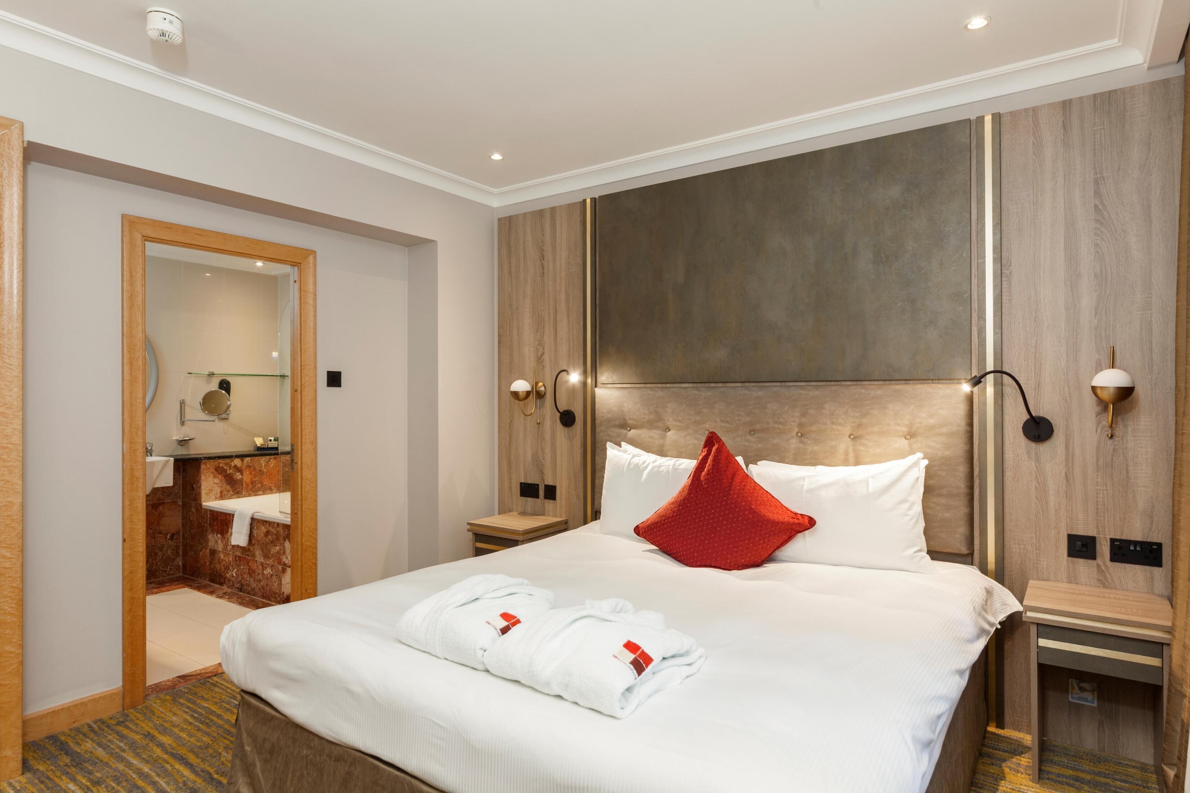A hotel room with a large bed, a wooden headboard, a mirror, a bathroom with a bathtub, and a red pillow.