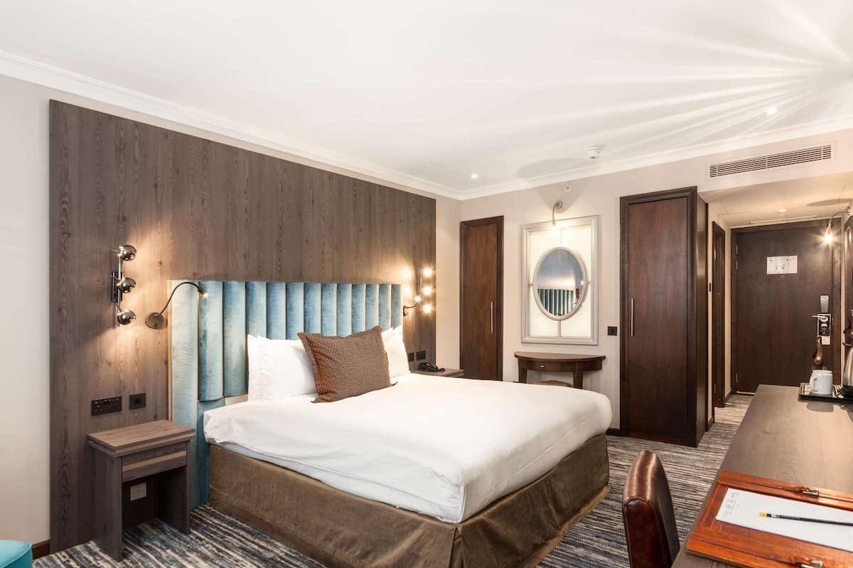 Ein Hotelzimmer mit einem großen Bett, einem Holz-Kopfteil, einem Nachttisch, einem Schreibtisch und einem Spiegel.