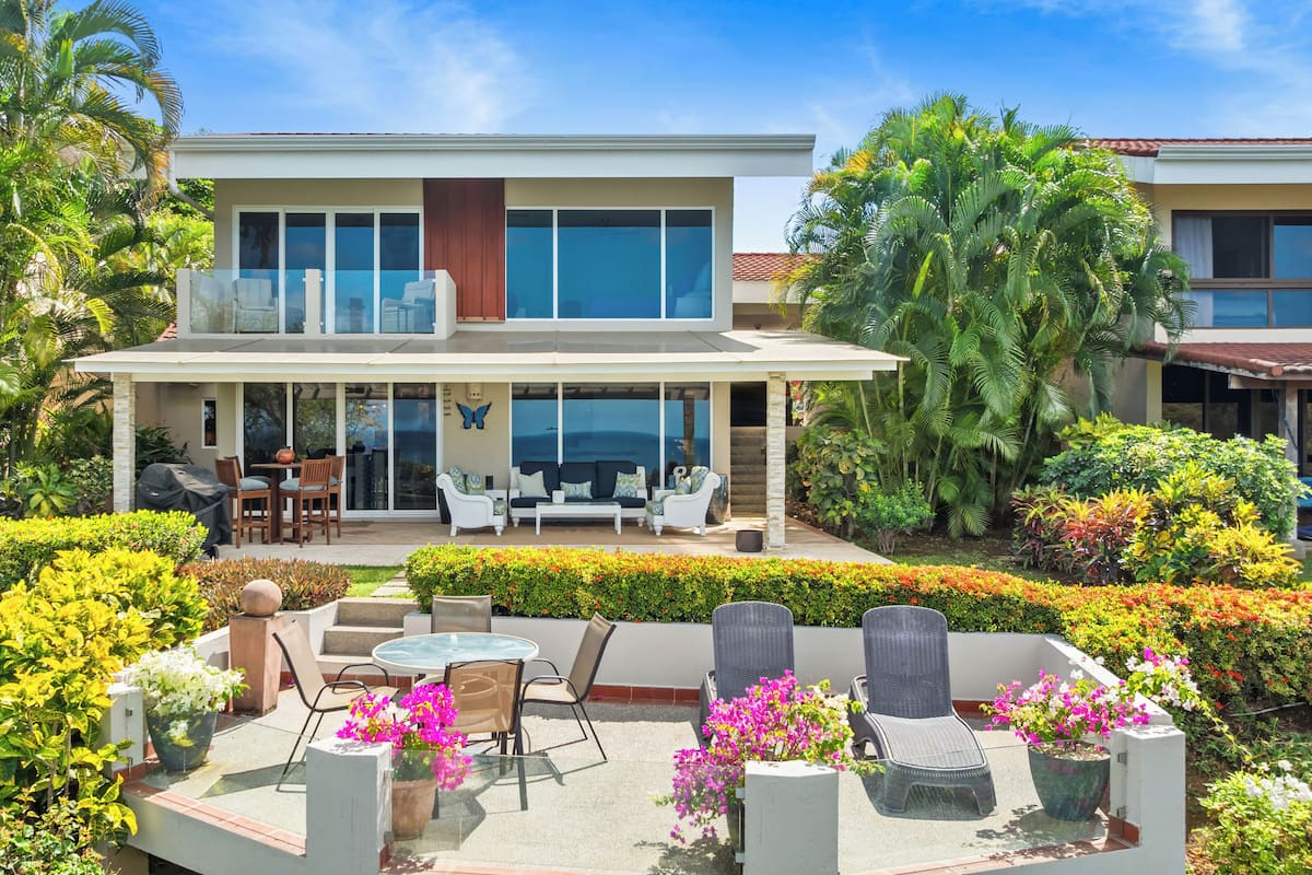 Ein modernes, zweistöckiges Haus mit überdachter Terrasse, Sitzgelegenheit im Freien und üppigem Grün.