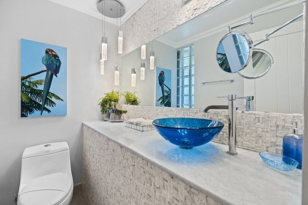 A modern bathroom with a white marble countertop, a blue glass bowl sink, and a white toilet. There are two wall-mounted mirrors, a potted plant, and a painting of a bird on the wall.