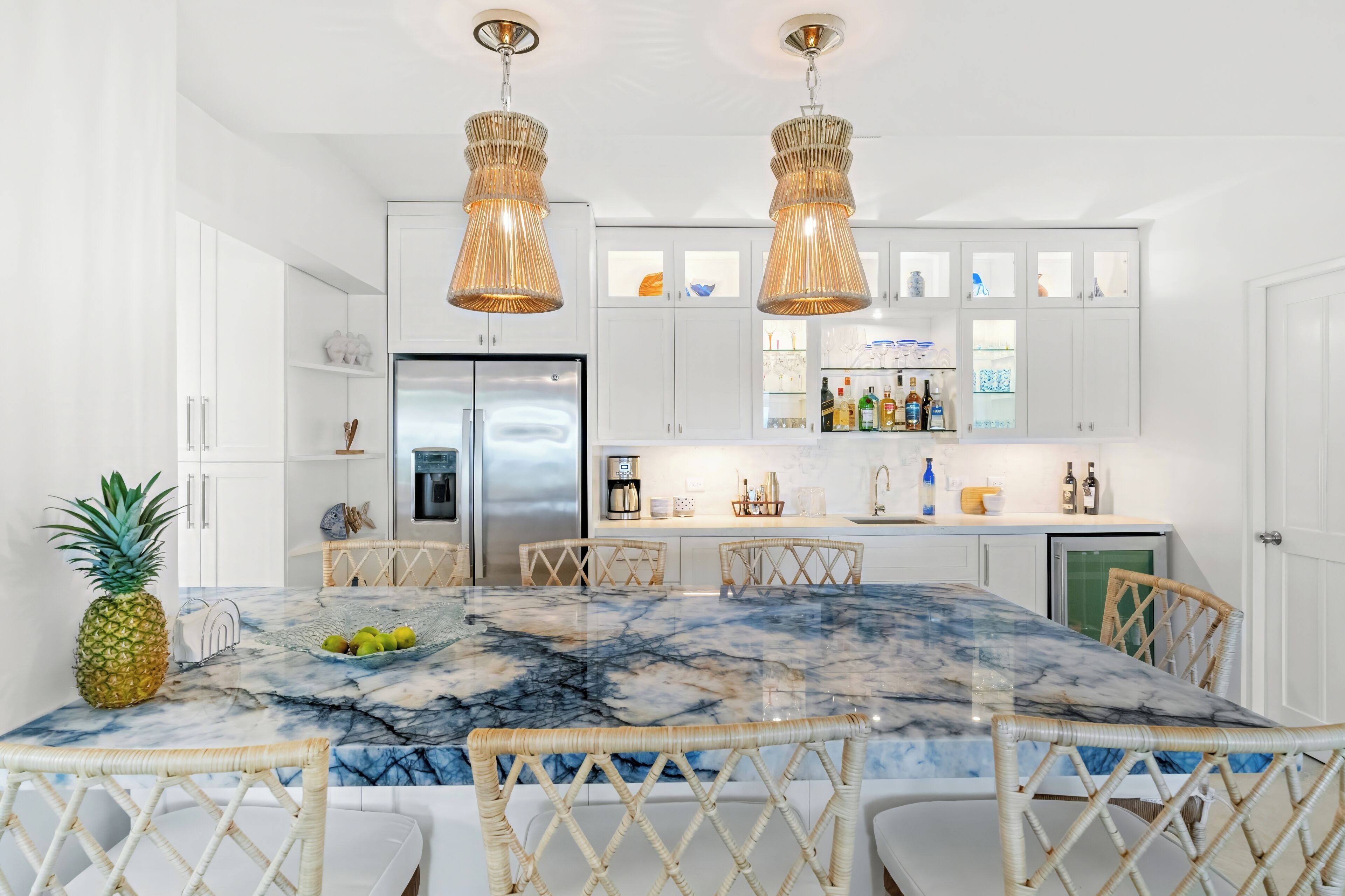 Une cuisine moderne avec des armoires blanches, un îlot en marbre, des chaises en osier et des luminaires suspendus.