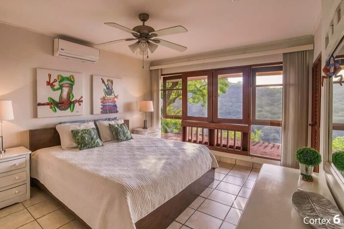 Ein Schlafzimmer mit einem Bett, Nachttischen, einem Deckenventilator und einem großen Fenster mit Blick auf Grünflächen.