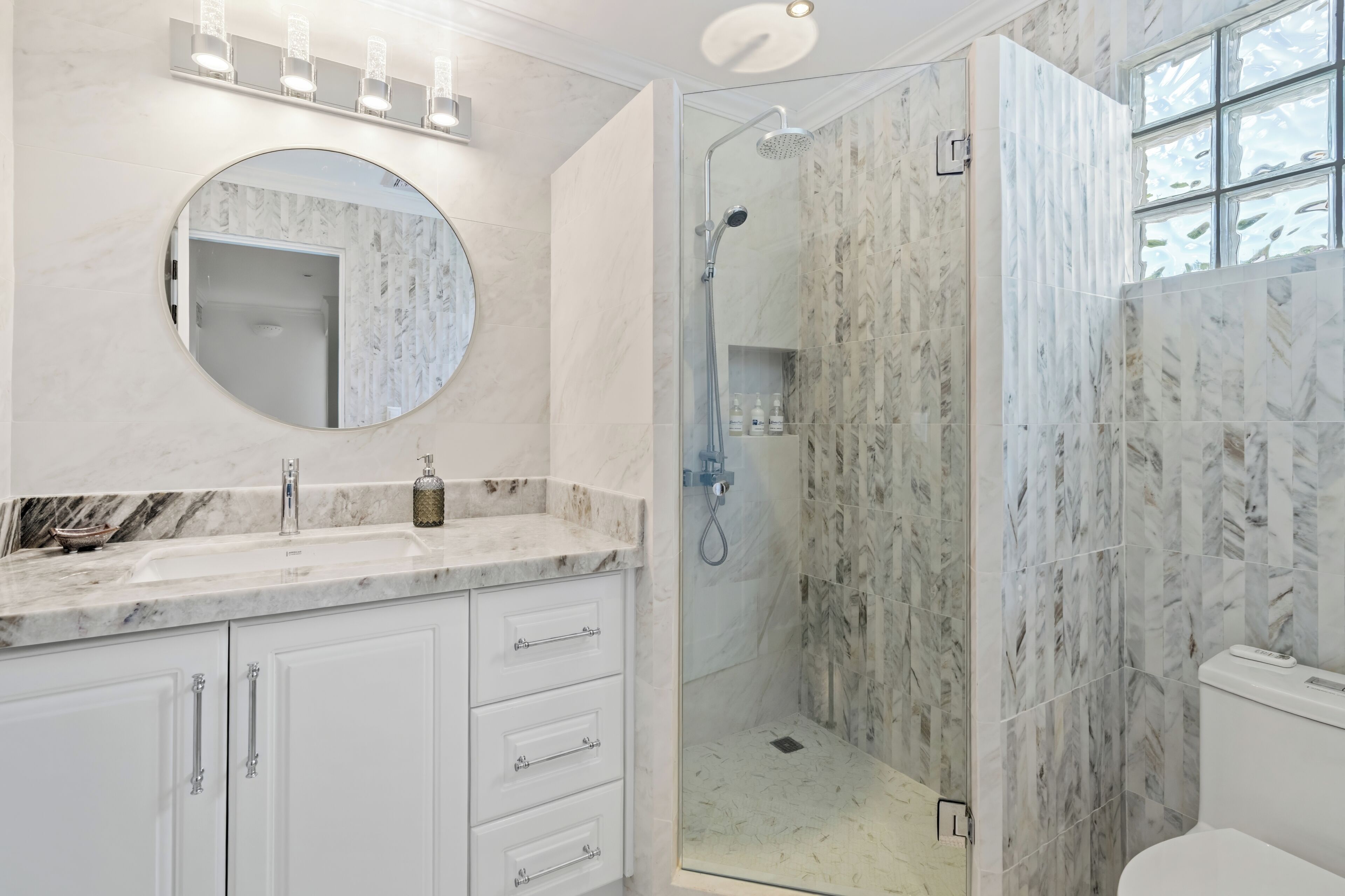 A bathroom with a marble sink, a round mirror, a glass-enclosed shower, and a toilet.