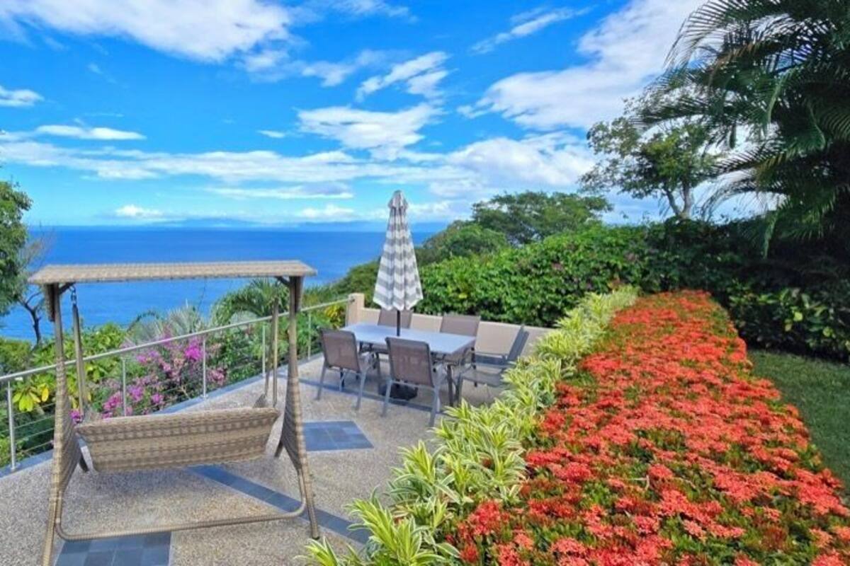 Eine Terrasse mit einer Schaukel, einem Tisch und Stühlen mit Blick auf die Küste und üppiges Grün.